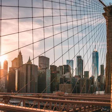The skyline of New York City with the Brooklyn Bridge in the foreground and the setting sun behind.