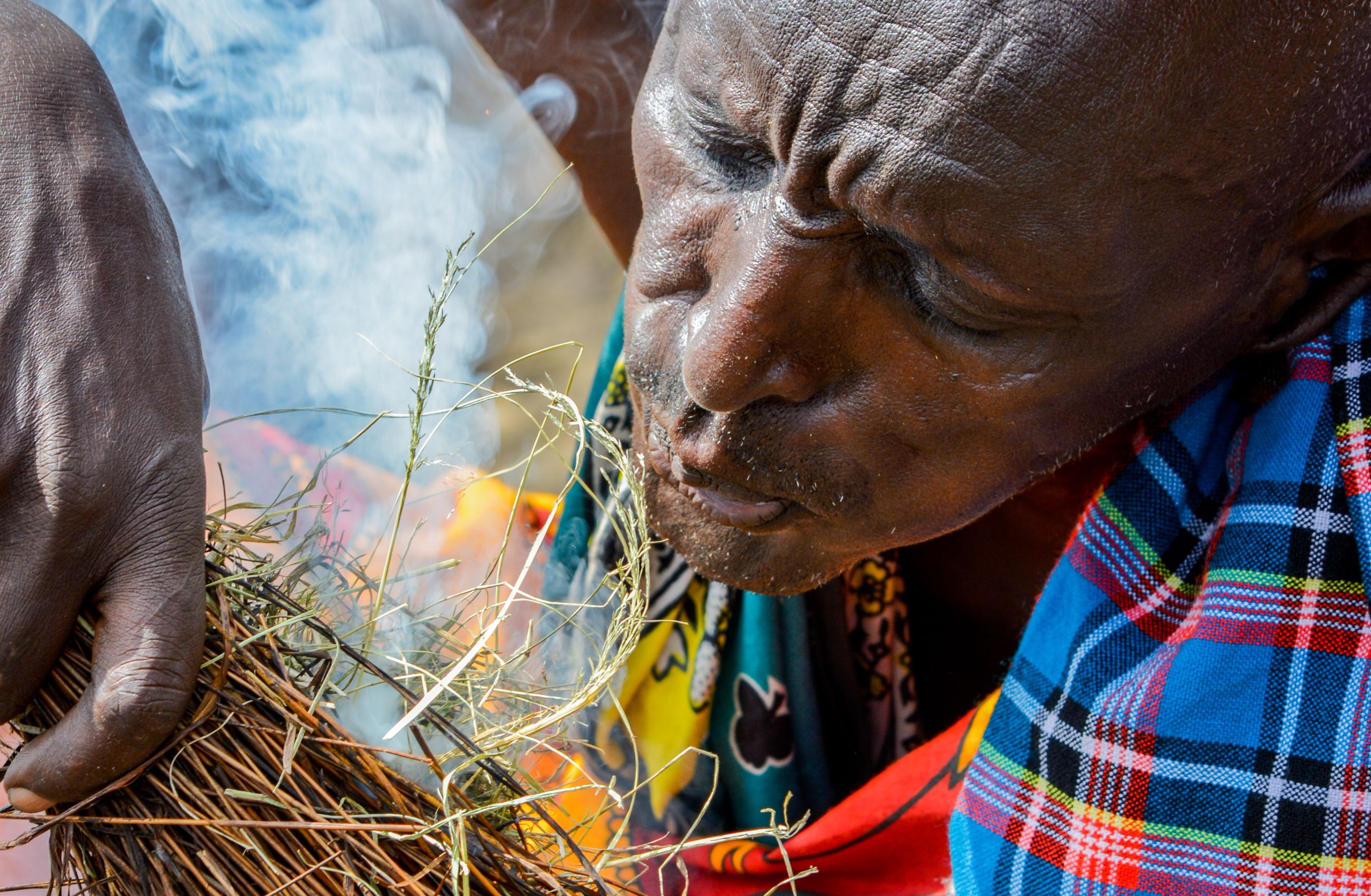 A man from a Maasai village in Kenya blows on grass kindling to feed a fire and smoke blows around his face.