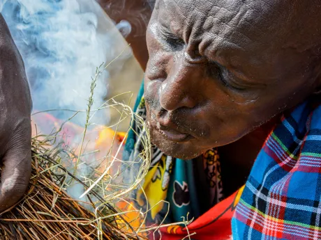 A man from a Maasai village in Kenya blows on grass kindling to feed a fire and smoke blows around his face.