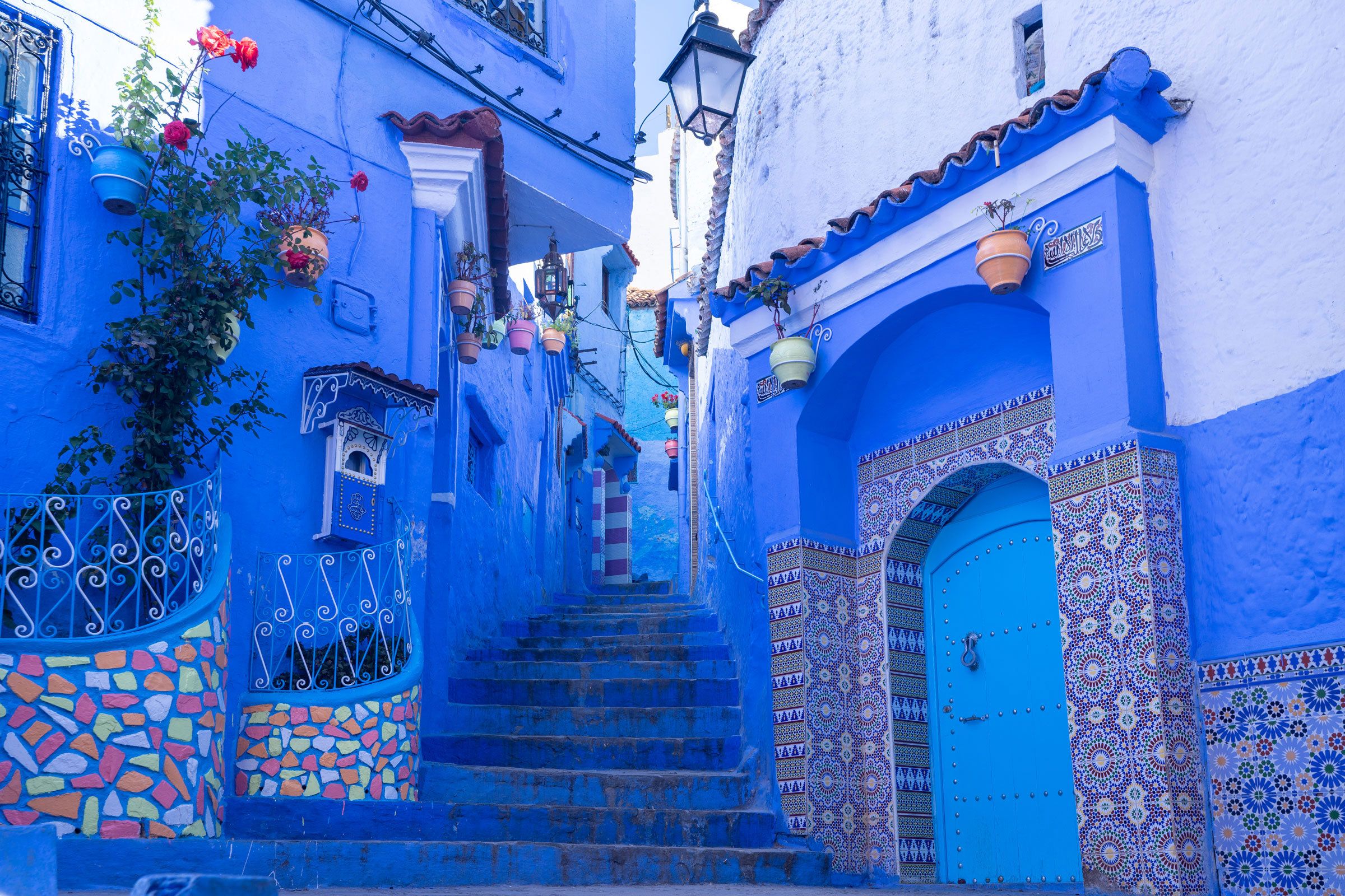 Blue-painted alley with tiled doorway, steps and hanging flower pots in Chefchaouen, Morocco