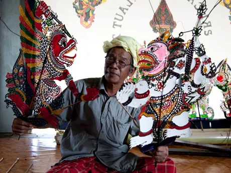 A man smiling at the camera with ornate puppets in Wayang Kulit Indonesia