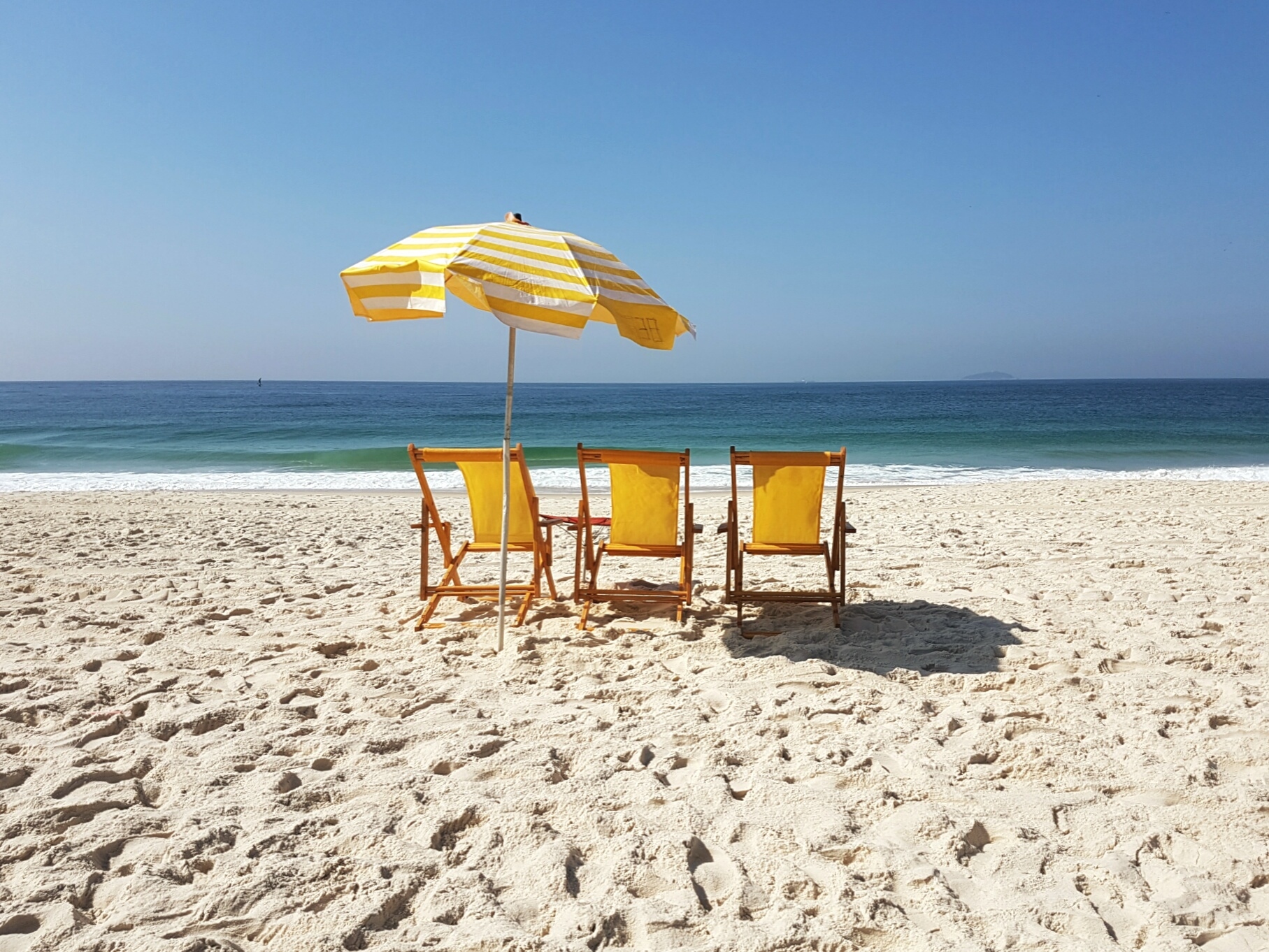 3 yellow chairs underneath an umbrella on a sandy, sunny beach