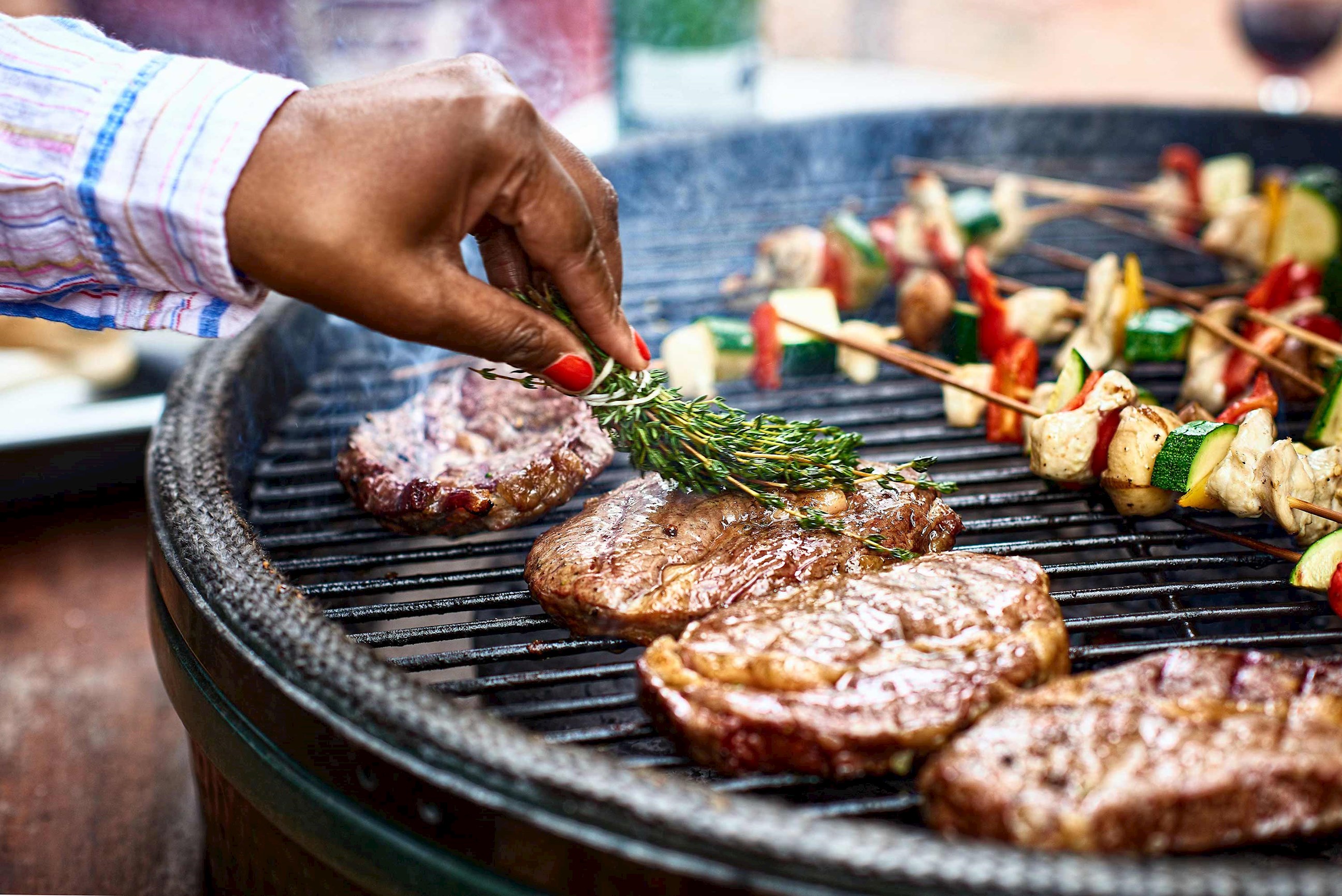Close-up of hand basting steaks with herbs on grill beside vegetables kebabs