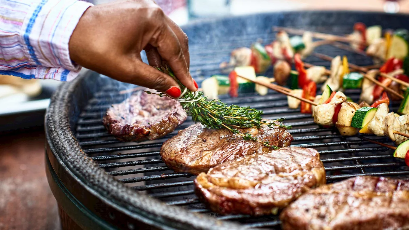 Close-up of hand basting steaks with herbs on grill beside vegetables kebabs
