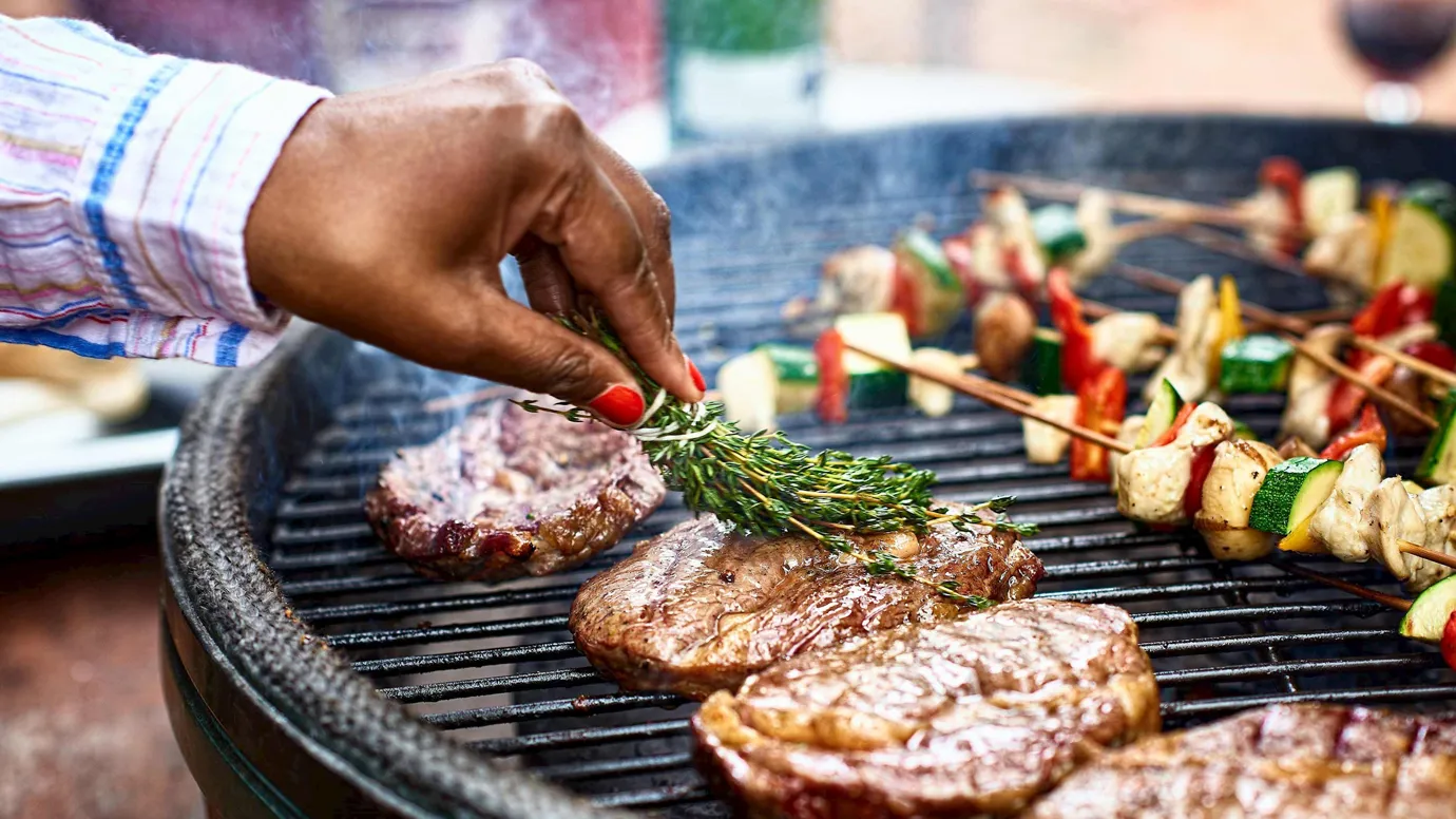 Close-up of hand basting steaks with herbs on grill beside vegetables kebabs