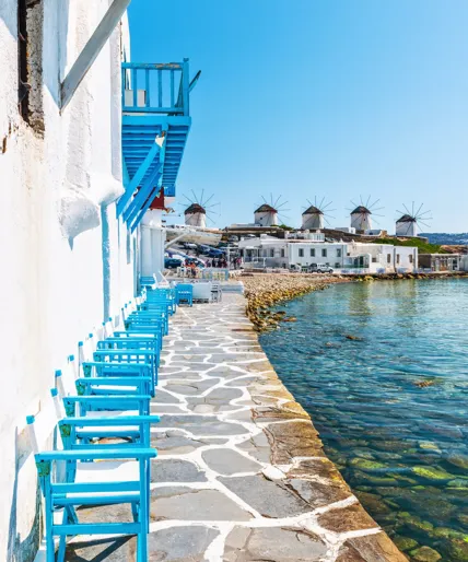 Looking down the seafront of a Greek harbour town on Mykonos with little blue chairs sitting on sunbleached flagstones.