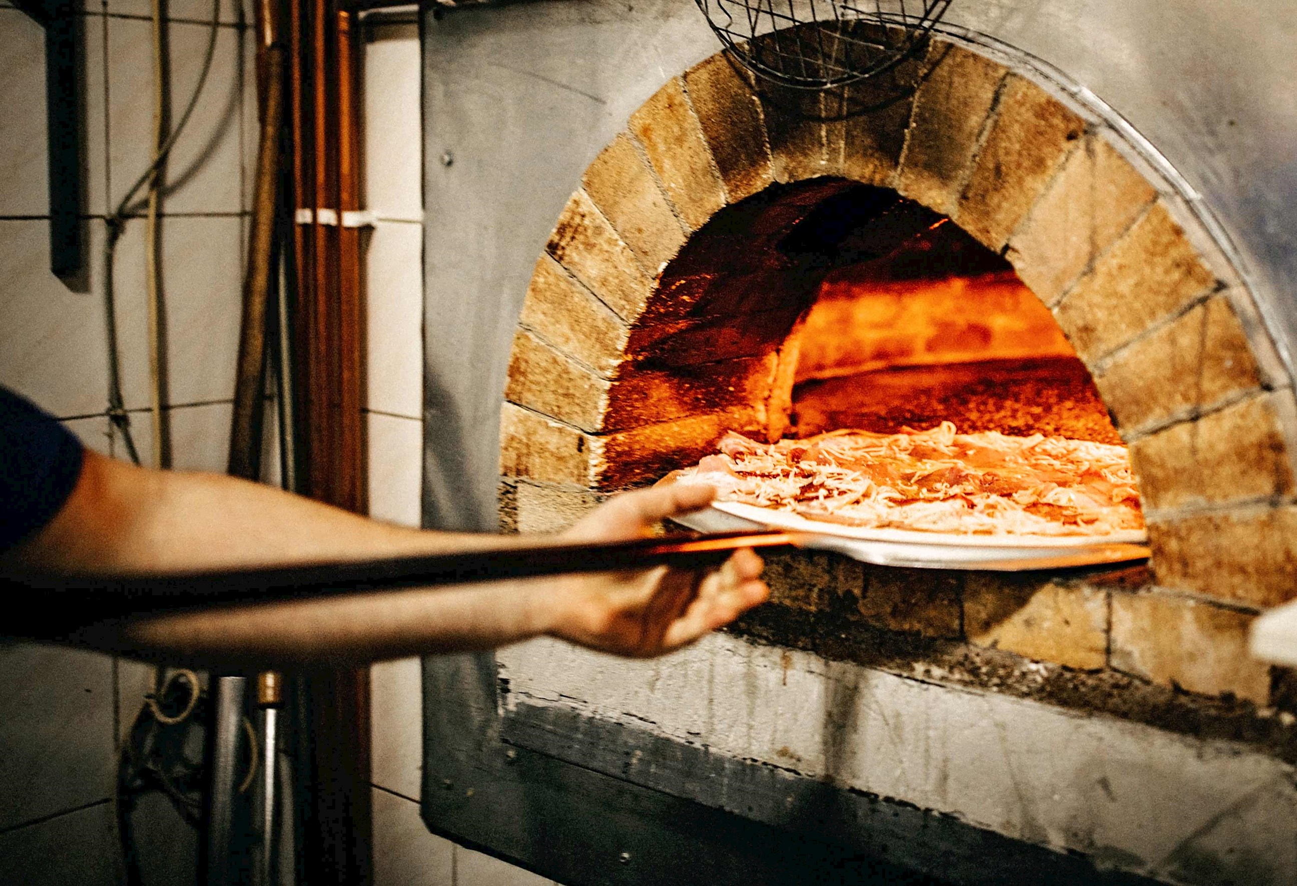 Pizza being placed into traditional wood-fired brick oven with glowing embers 