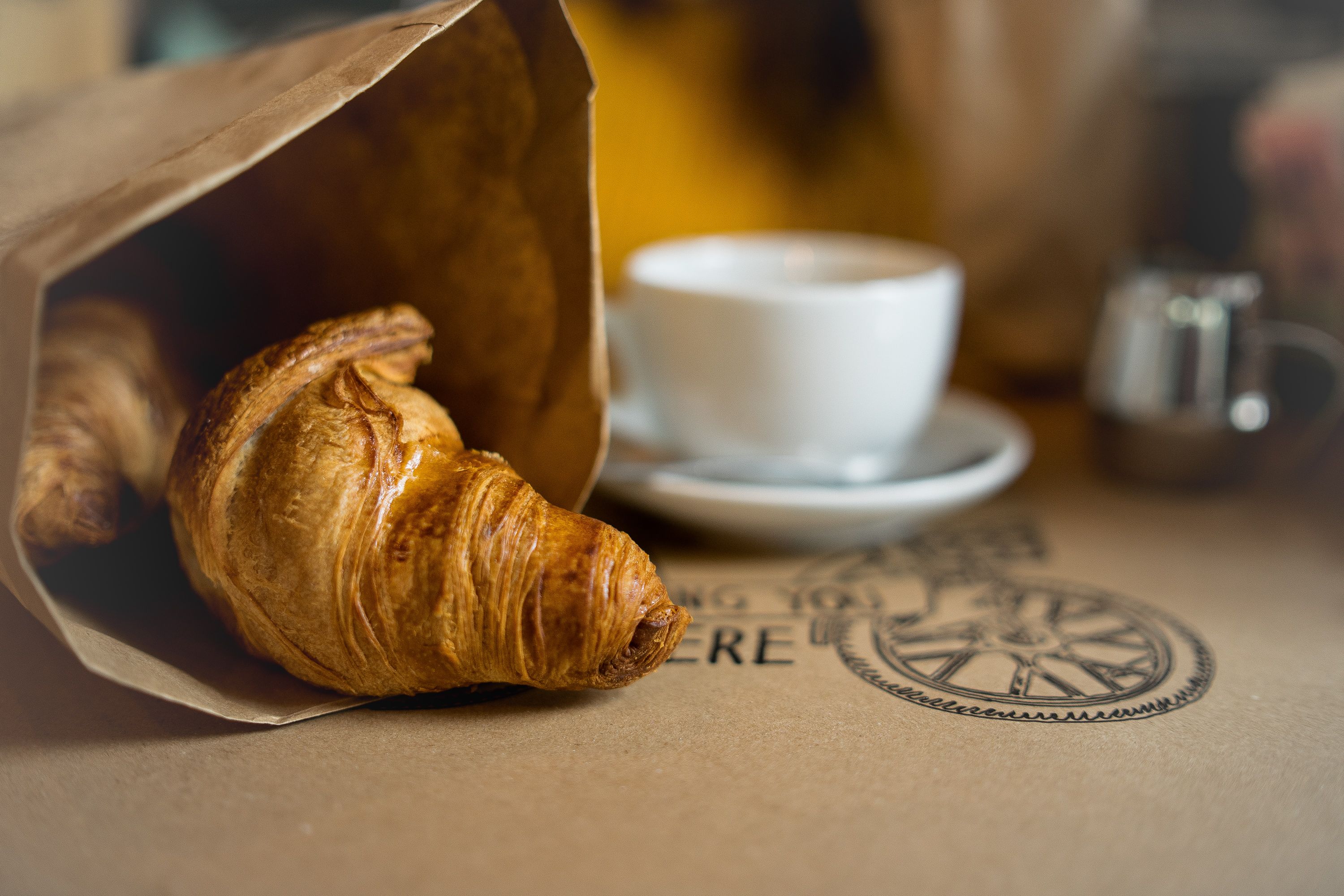 A croissant half poking out of a brown paper bag with a small cup of coffee. A common food choice in France.