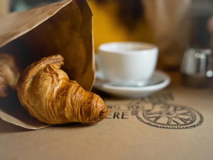 A croissant half poking out of a brown paper bag with a small cup of coffee. A common food choice in France.