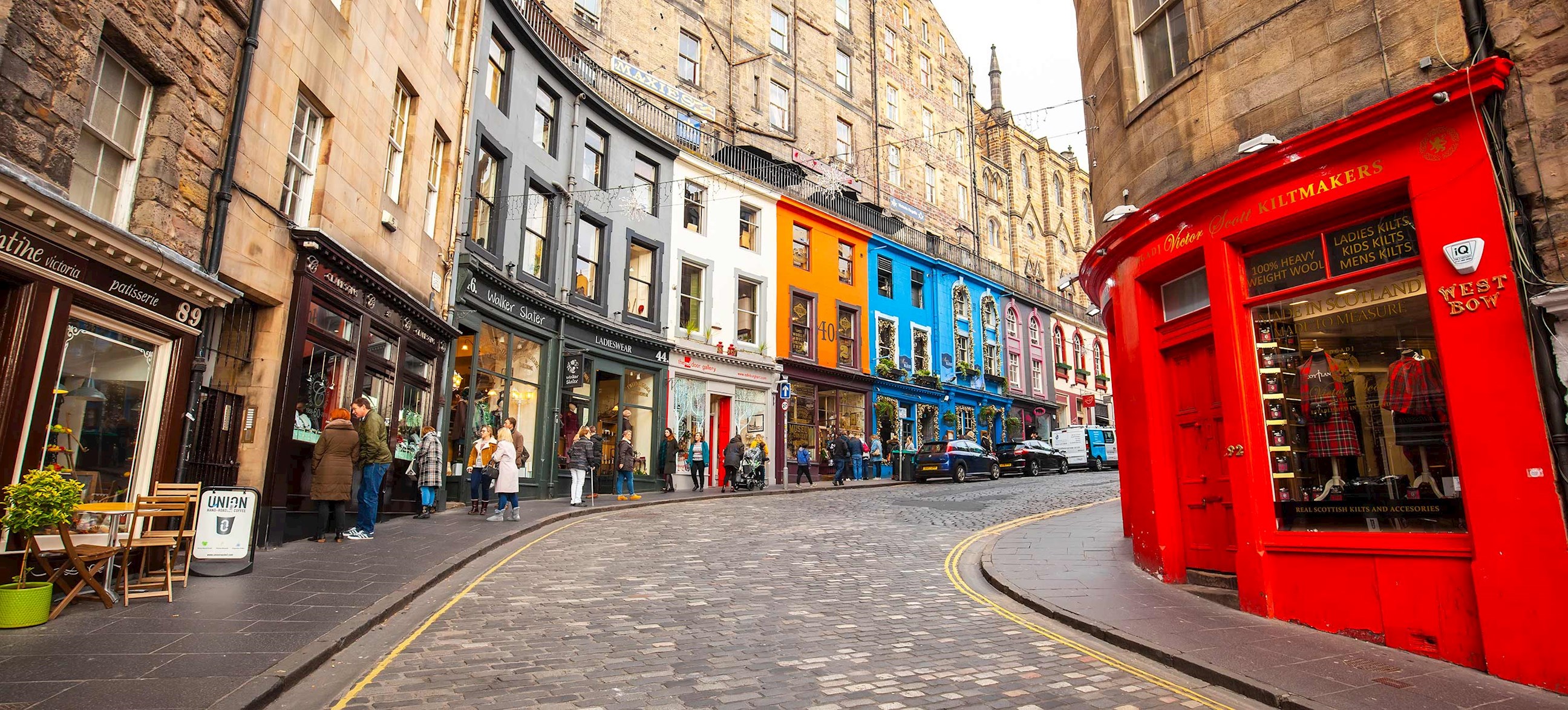Curved cobblestone street lined with colourful shopfronts and red building in Edinburgh, Scotland