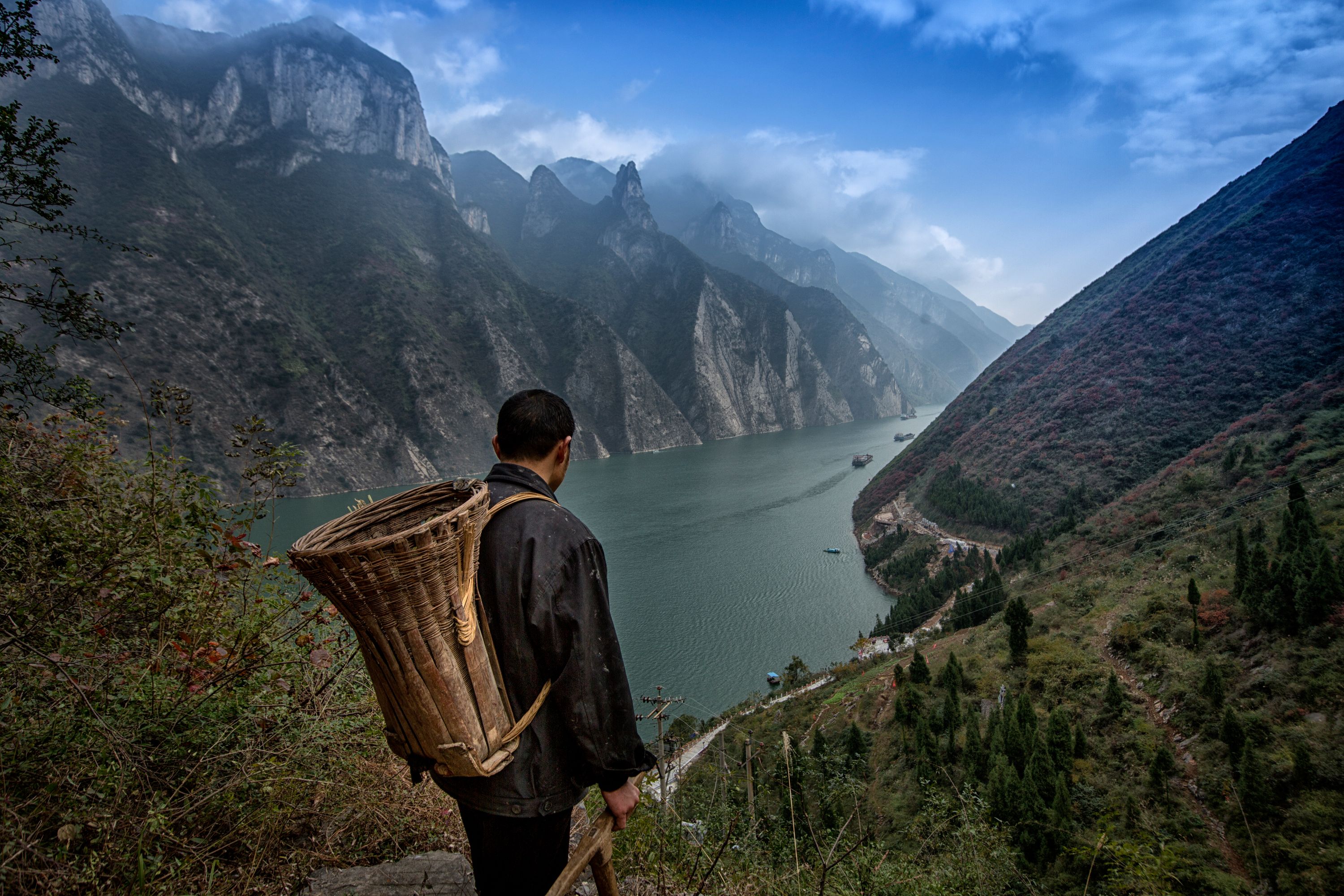 A man carrying a traditional basket on his back looks out at the Yangtze River in China as it passes through a steep gorge.