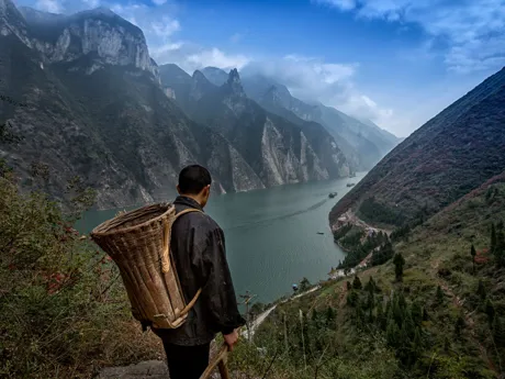 A man carrying a traditional basket on his back looks out at the Yangtze River in China as it passes through a steep gorge.