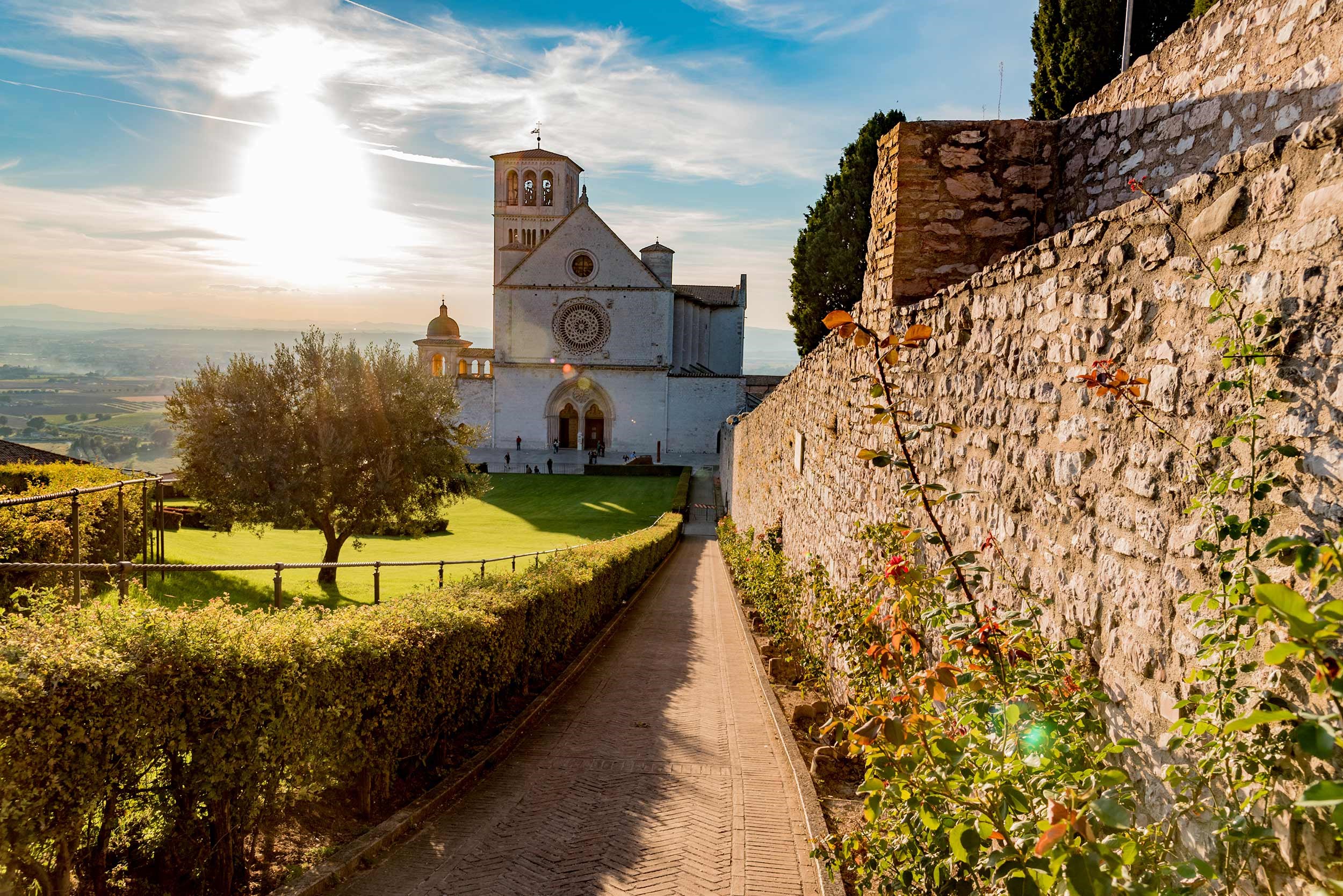 A stone pathway leading to a building with a rose window at sunset in Assisi, Italy